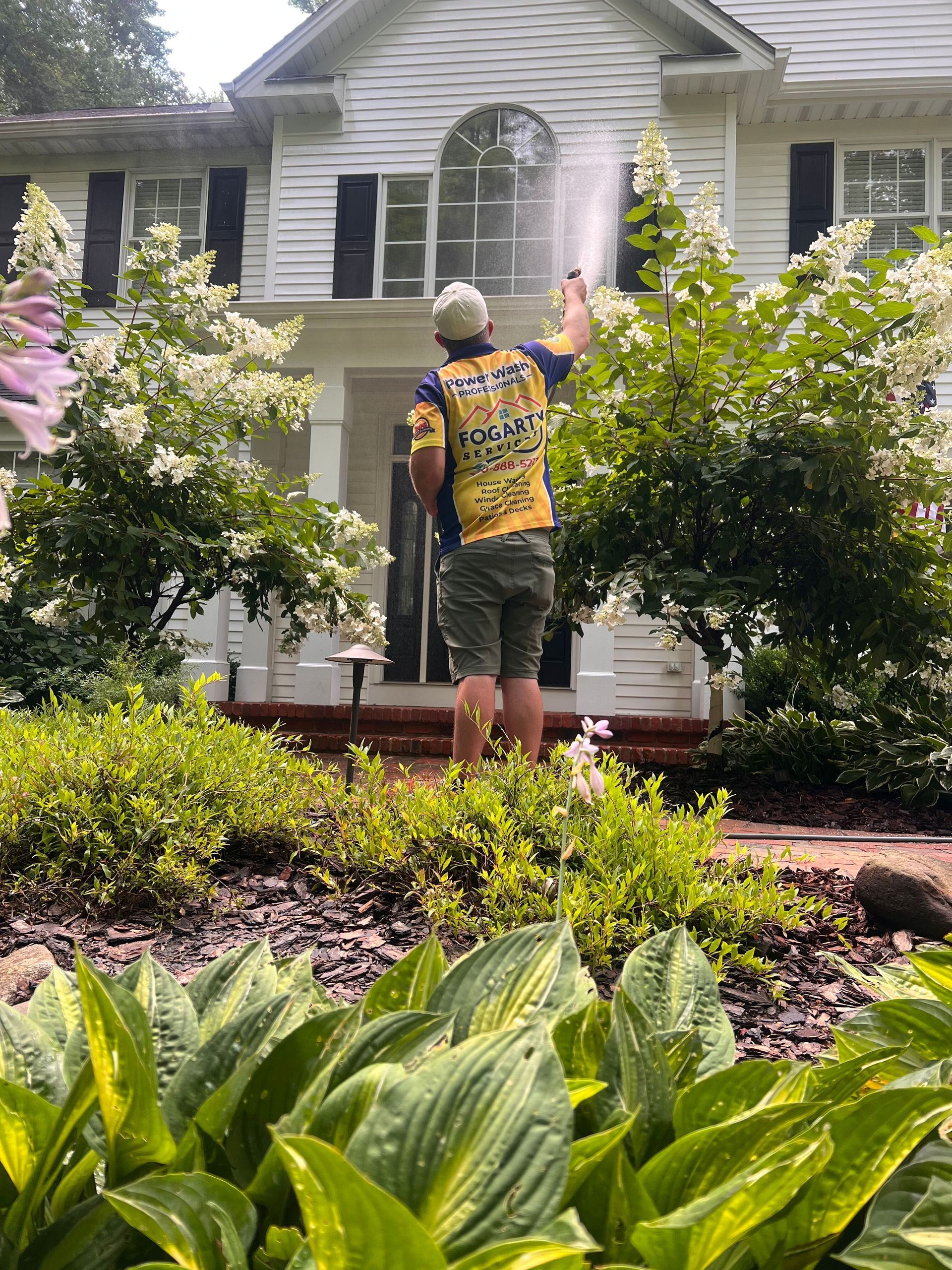 A person wearing a yellow jersey power washes the exterior of a white, two-story house surrounded by flowering bushes.