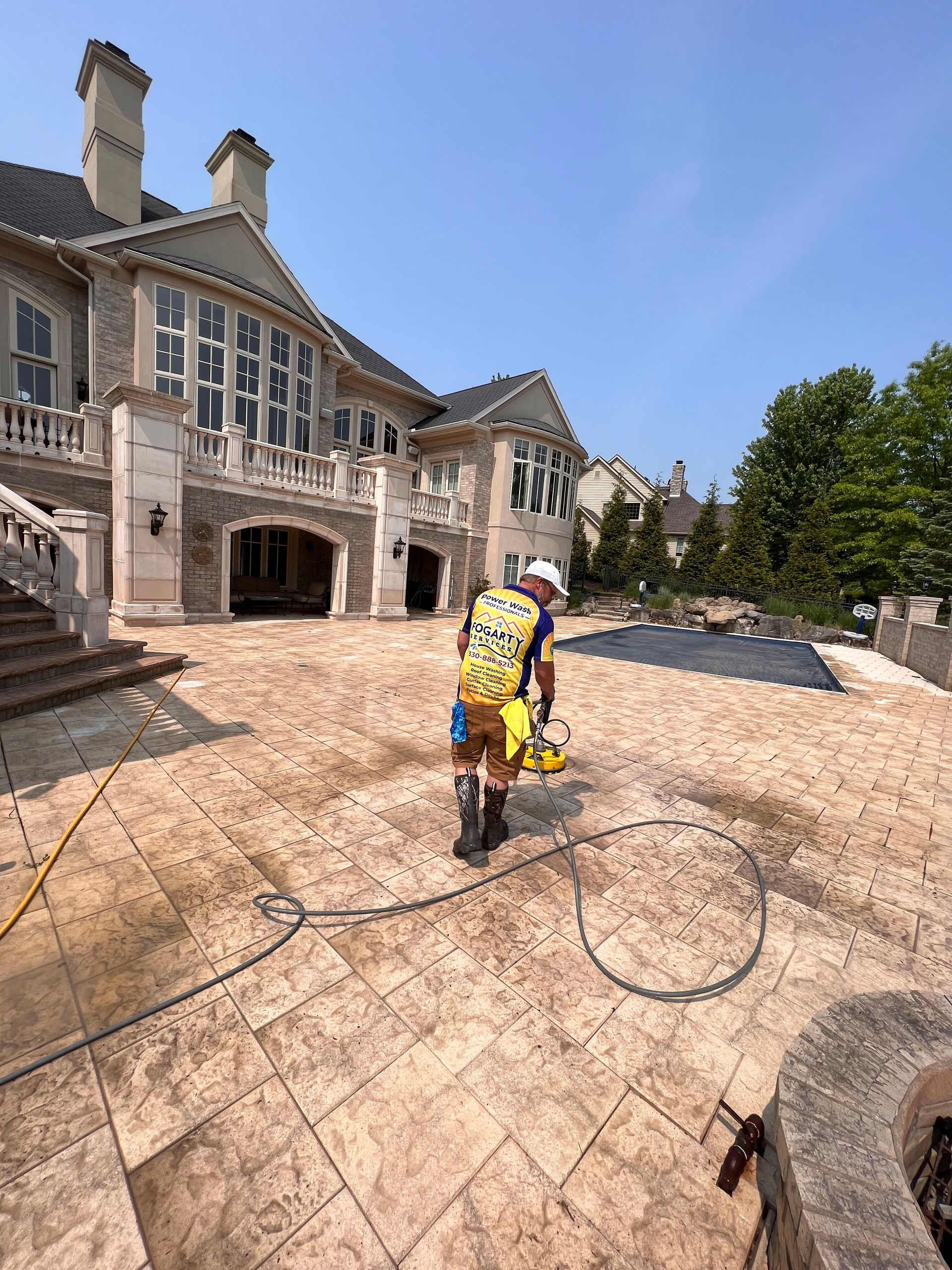 A worker wearing a bright yellow backpack uses a surface cleaner on a large stone patio behind a multi-story mansion.