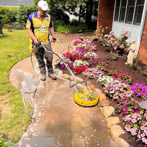 A worker uses a circular power washer to clean a brick walkway beside a garden bed filled with colorful blooming flowers.