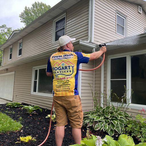 A person in a yellow and blue uniform power washing the exterior of a tan, two-story house.