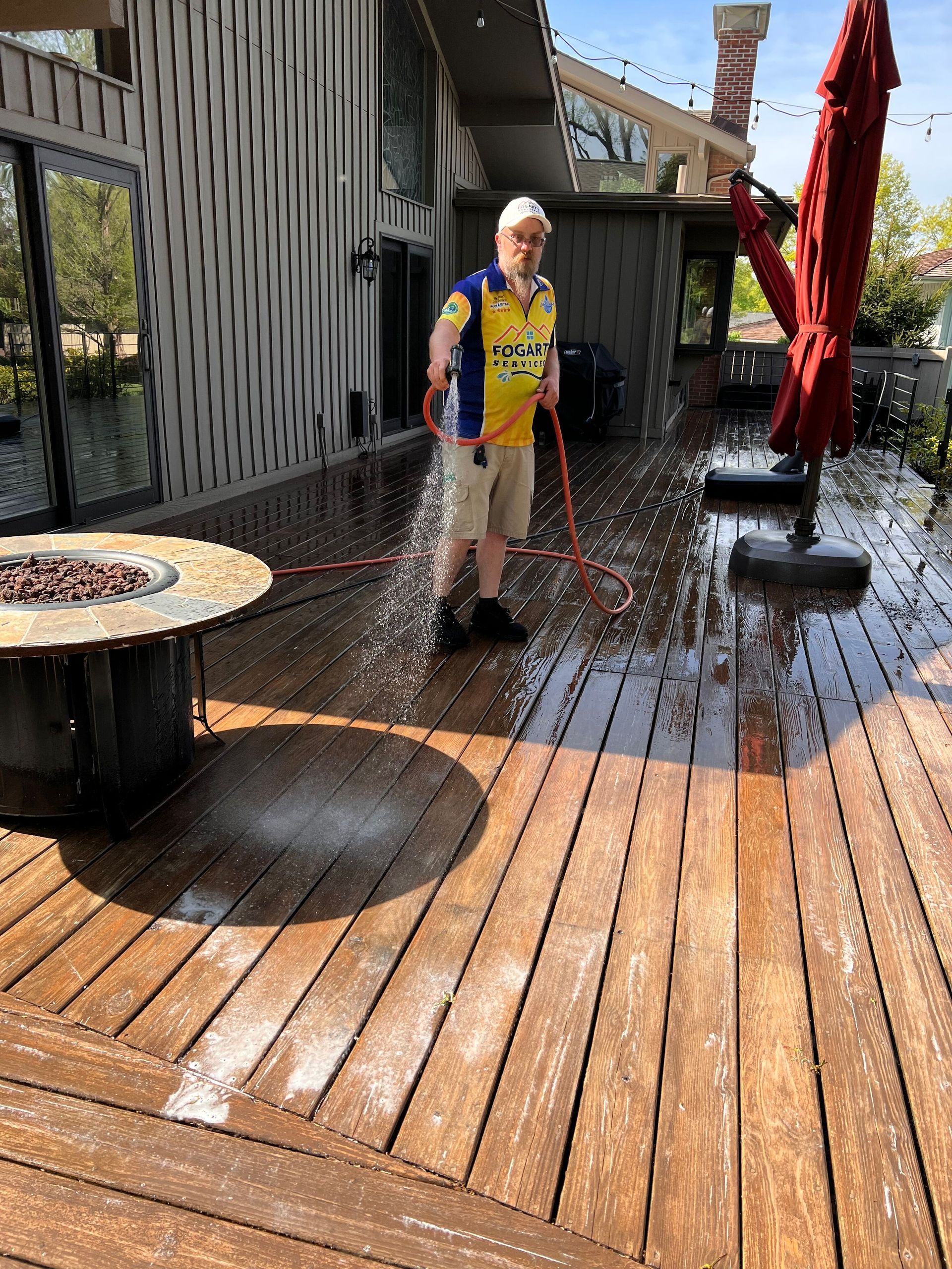 A person in a yellow and blue shirt uses a hose to spray water onto a wooden deck next to a fire pit and red umbrella.