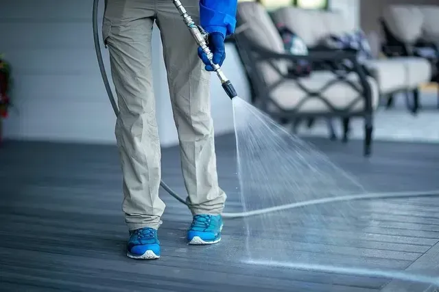 A person in khaki pants and blue sneakers pressure-washes a grey deck on an outdoor patio.