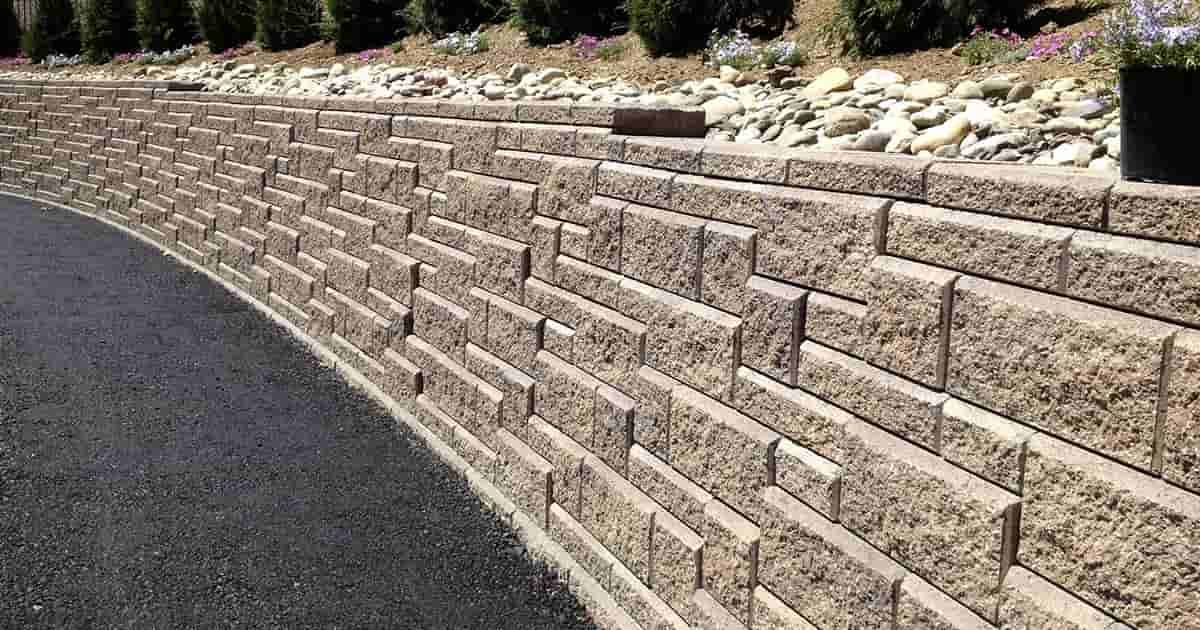 A curved retaining wall made of stacked, textured, tan stone blocks alongside an asphalt path with a rock-filled garden.