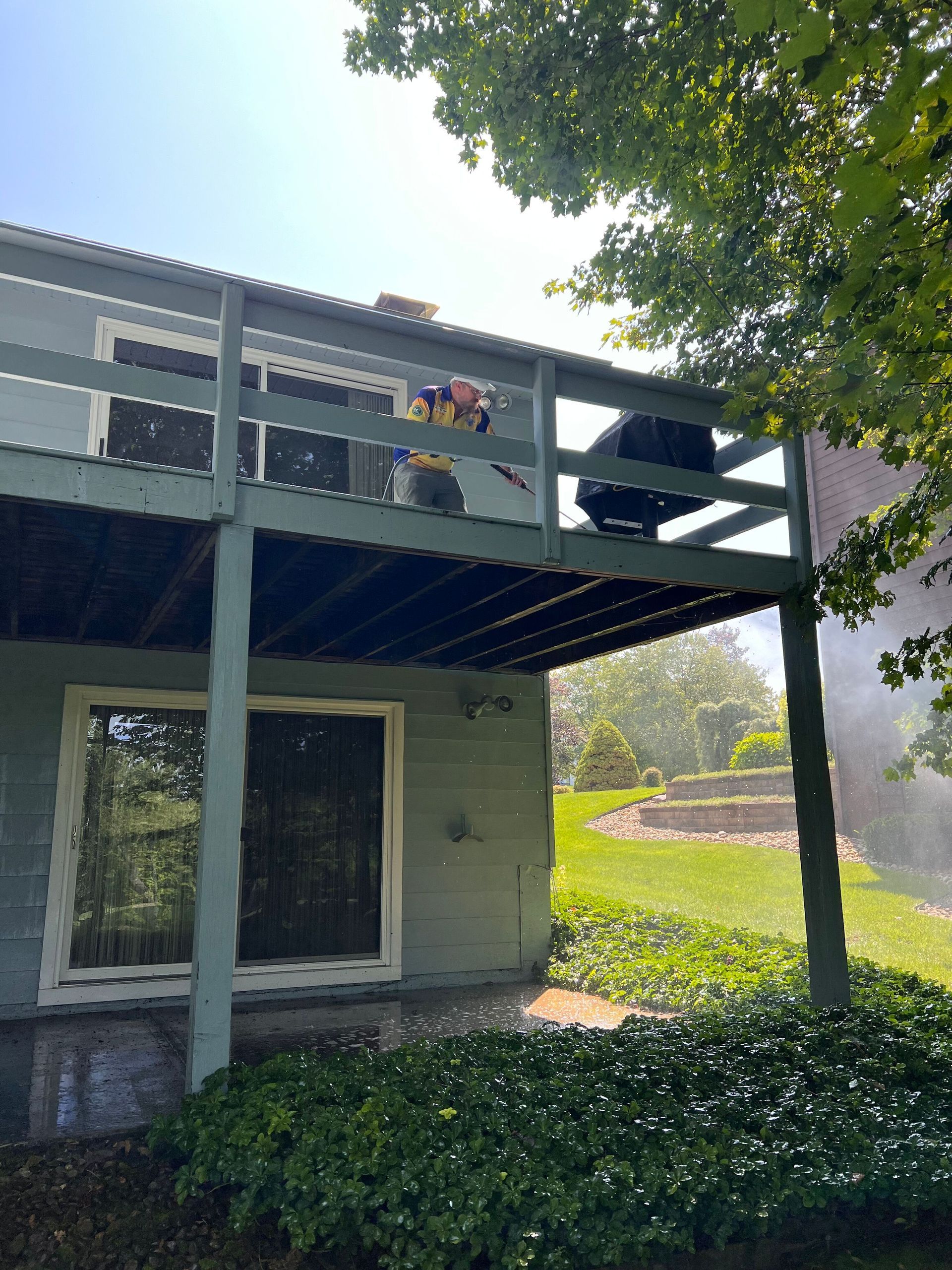 A person stands on a wooden deck attached to a gray house, appearing to operate a tool while smoke or mist is nearby.