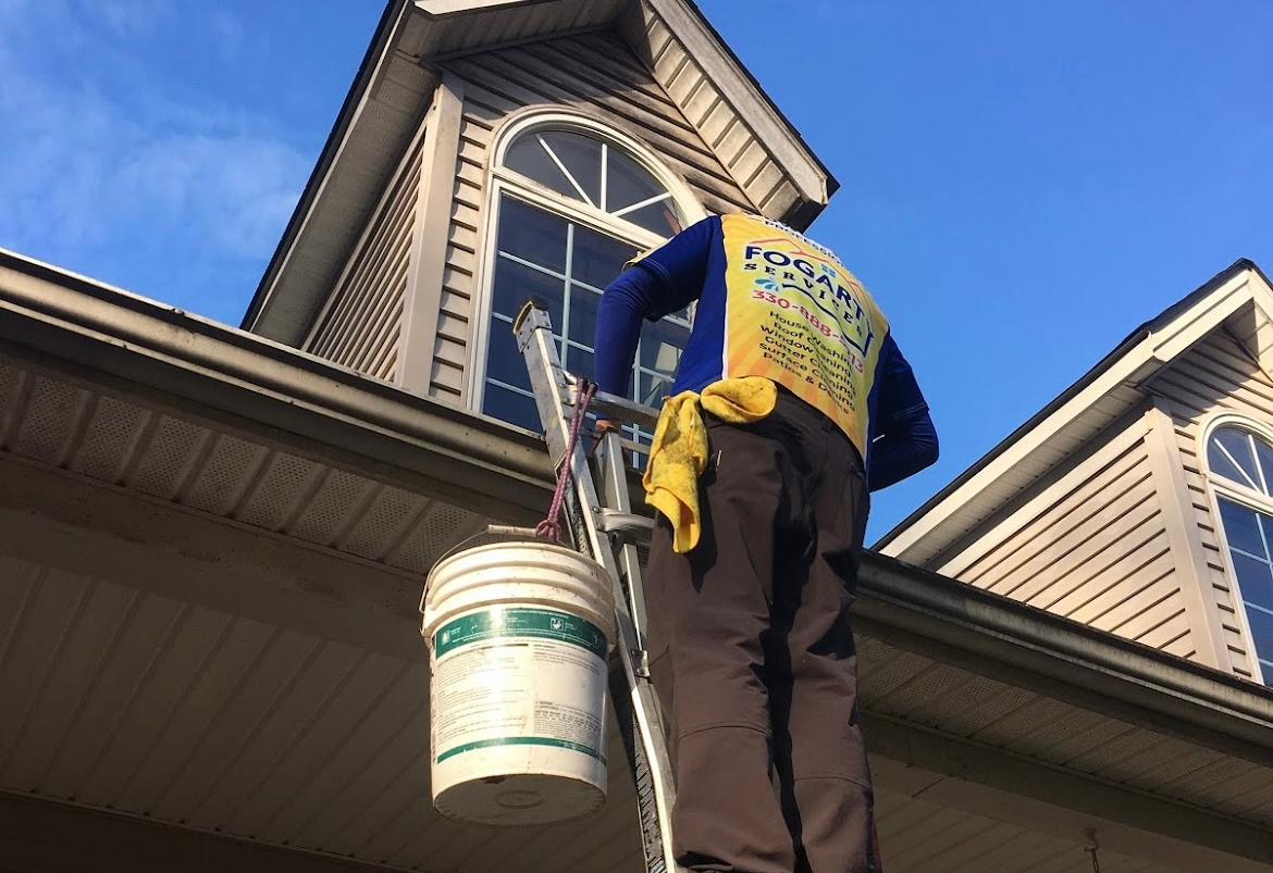 A worker in a yellow reflective vest stands on a ladder cleaning a home exterior with a bucket hanging from the ladder.