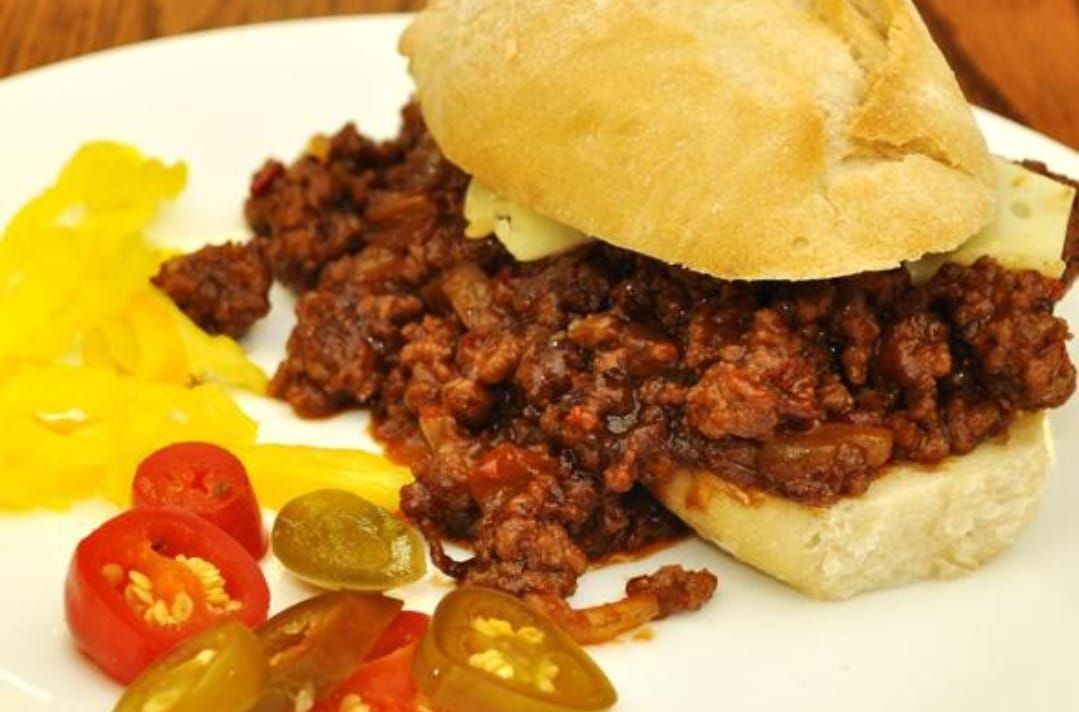 Panino Sloppy Joe con verdure sottaceto e contorno giallo su un piatto bianco.