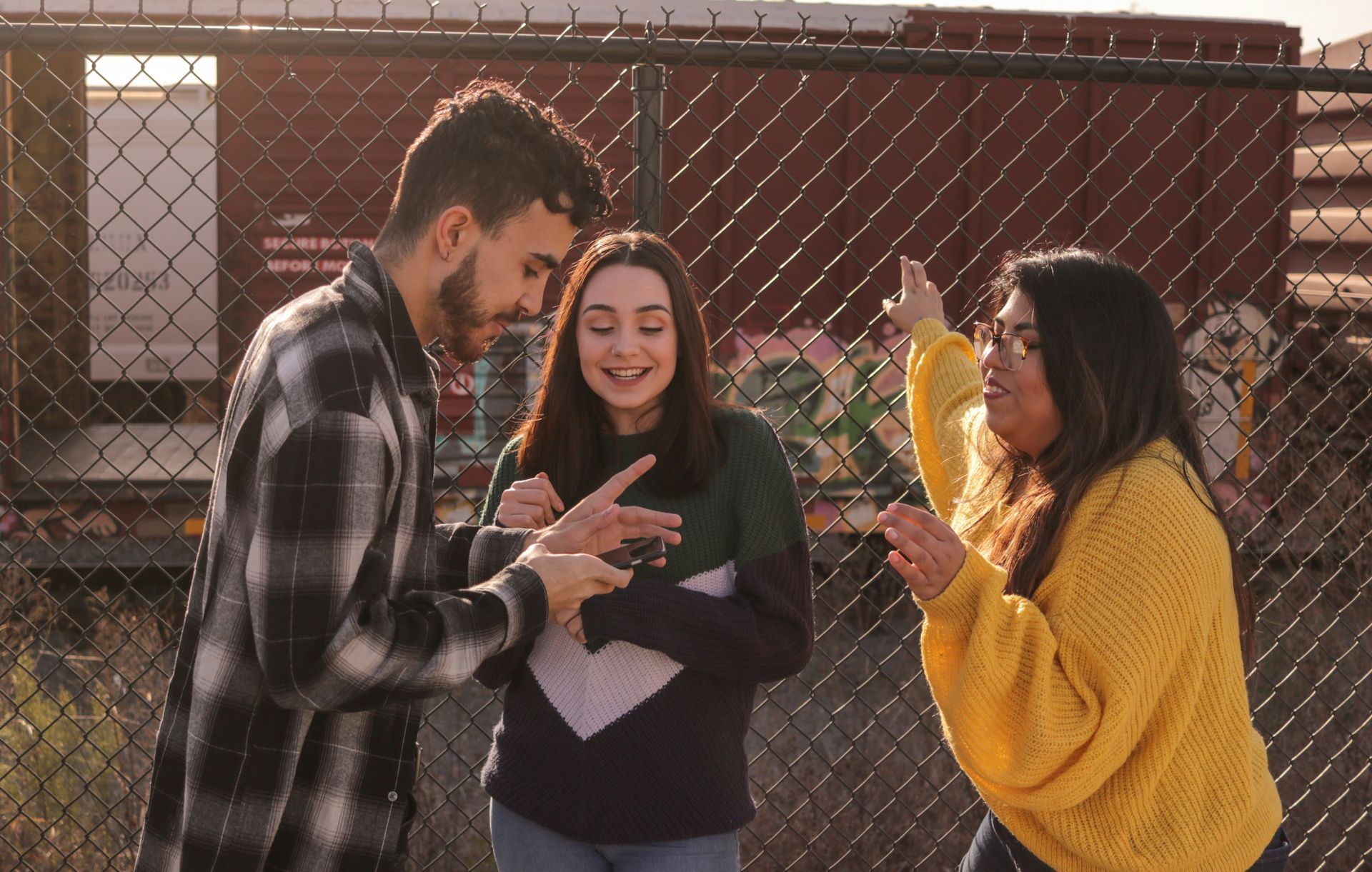 Three people stand by a chain-link fence, laughing and interacting with a small object while illuminated by warm light.