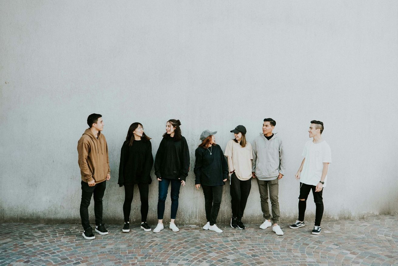 A group of seven people stand in a line against a light gray wall, facing each other and talking in casual clothing.
