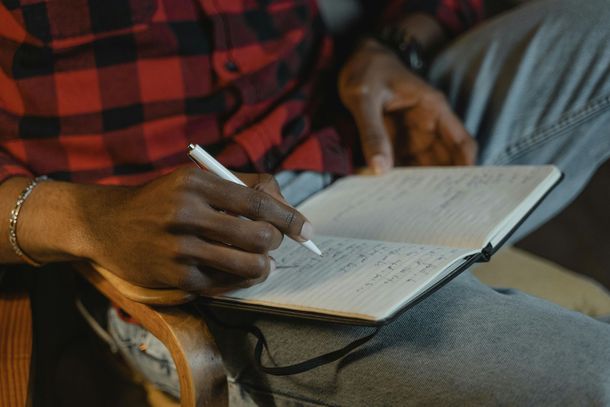 A person in a red plaid shirt writes in a lined notebook while sitting in a chair.