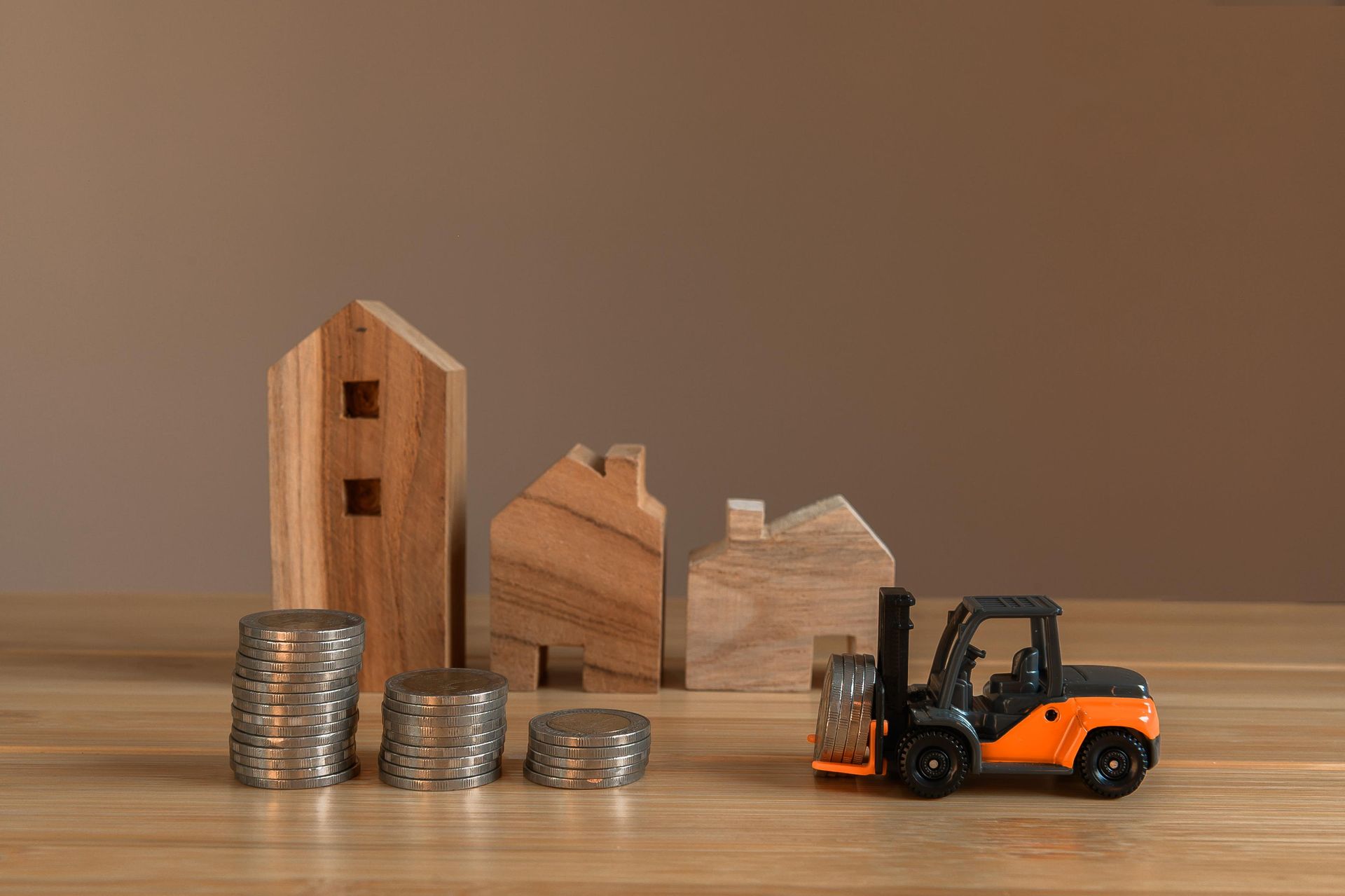 Stacks of coins, wooden house models, and a small orange forklift on a wooden table.