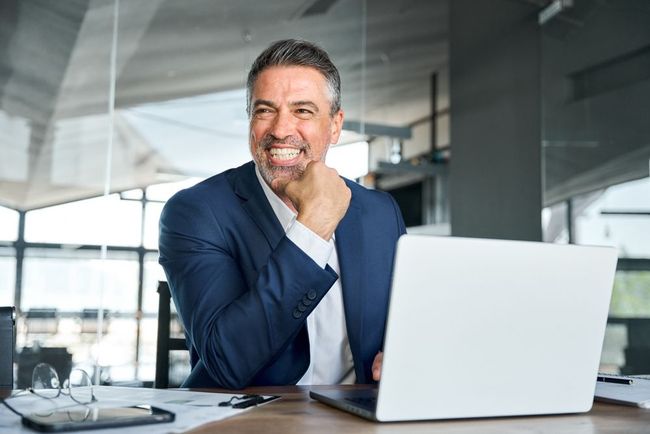 Smiling man in a navy blazer sitting at a desk with a laptop in a modern office