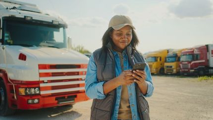 Woman in blue jacket using a phone in a truck lot, with colorful parked trucks behind her.