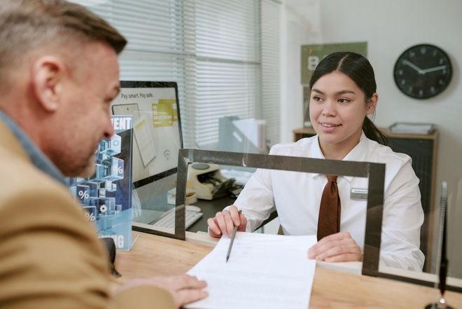 Two people exchange documents at a desk in an office, separated by a clear protective barrier.
