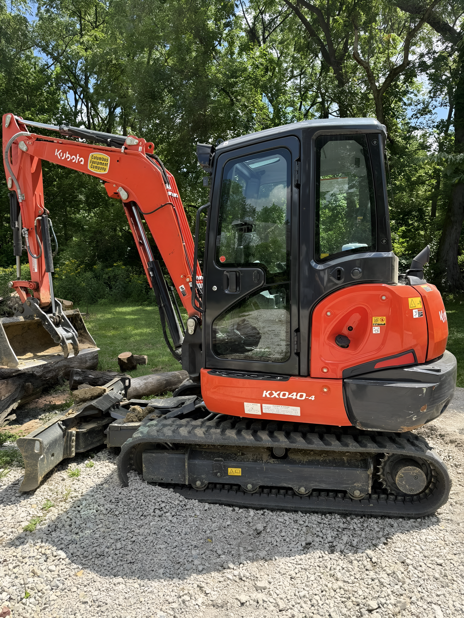 Orange Kubota excavator on gravel, cab with windows, bucket attachment, green trees in background.