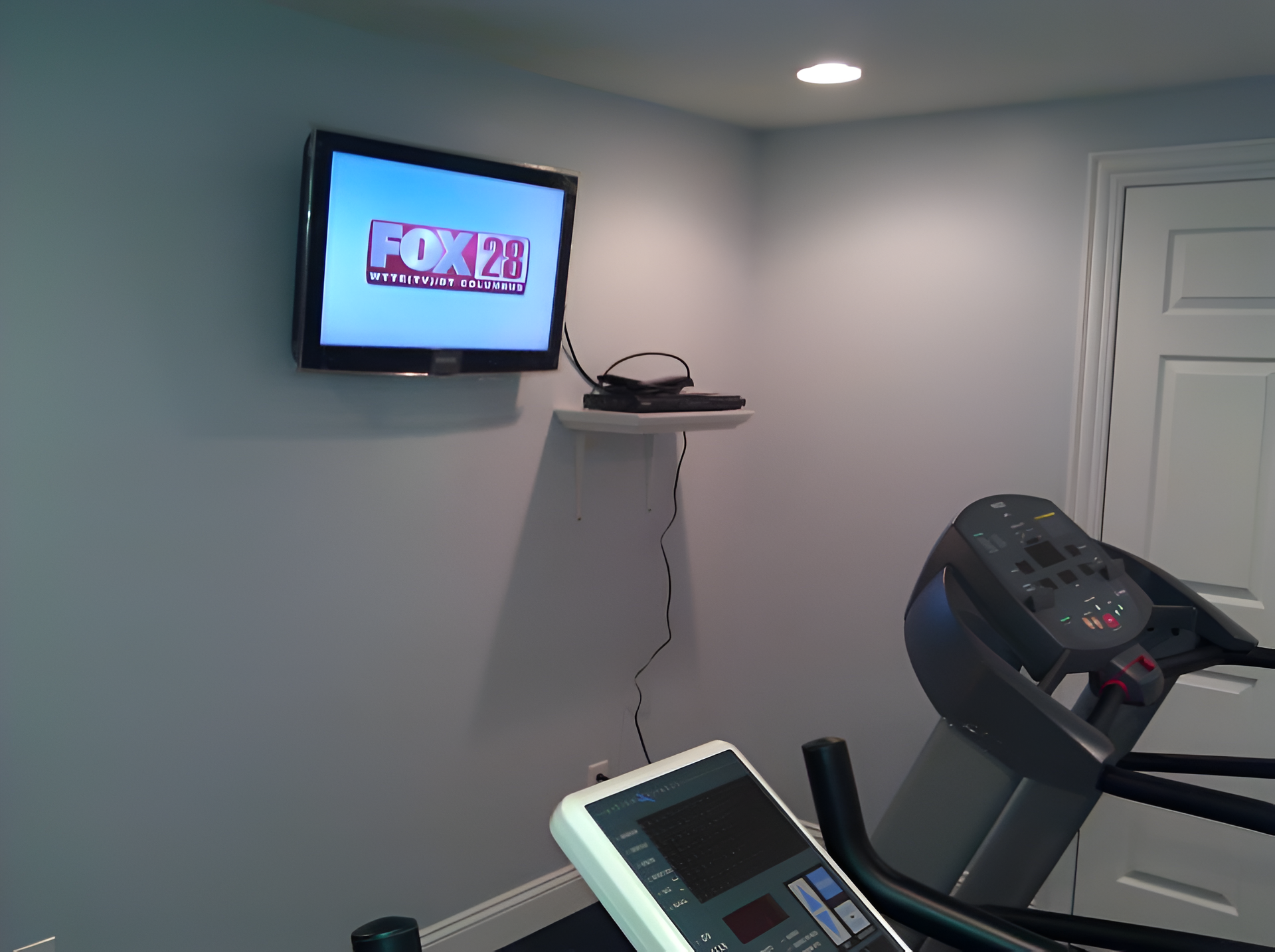 TV on wall above treadmill in a gym. Gray walls, white door, and shelf with electronic device.