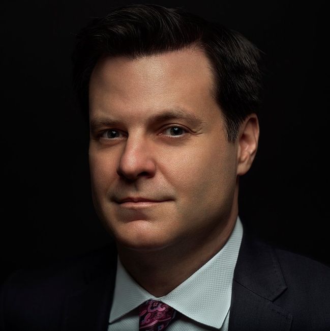 Portrait of a man in a dark suit and pink tie against a black background