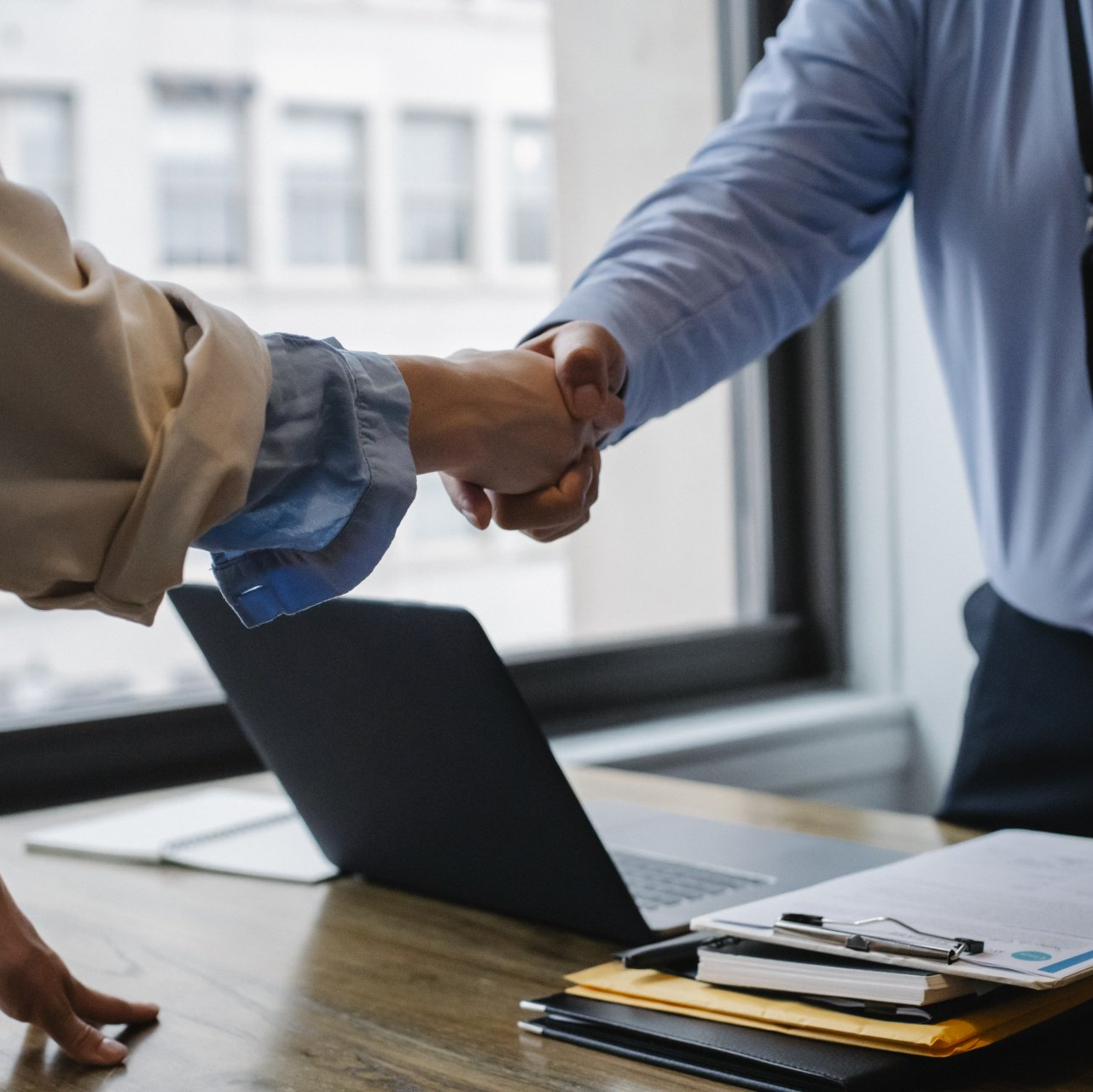 Two people shaking hands beside an open laptop and stack of papers in an office.