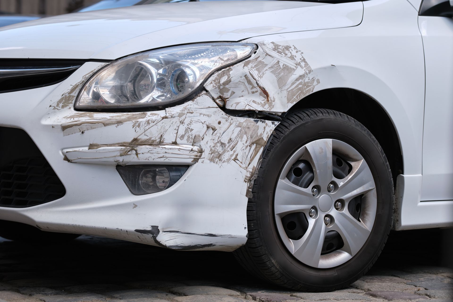 Front-left of a white car with heavy dirt and damage around the headlight and bumper
