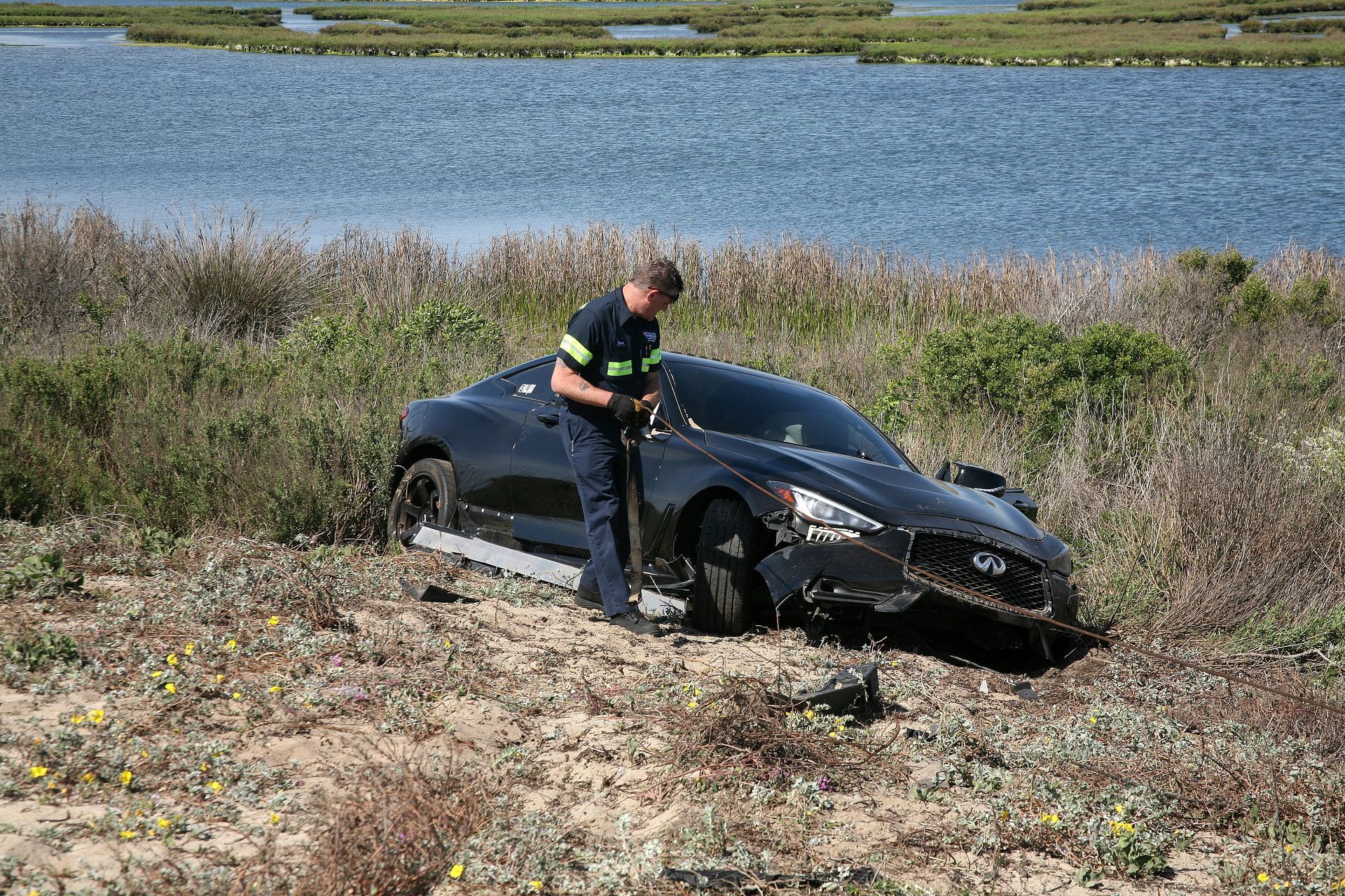 Police officer inspects a wrecked black car beside a lake in a grassy field.