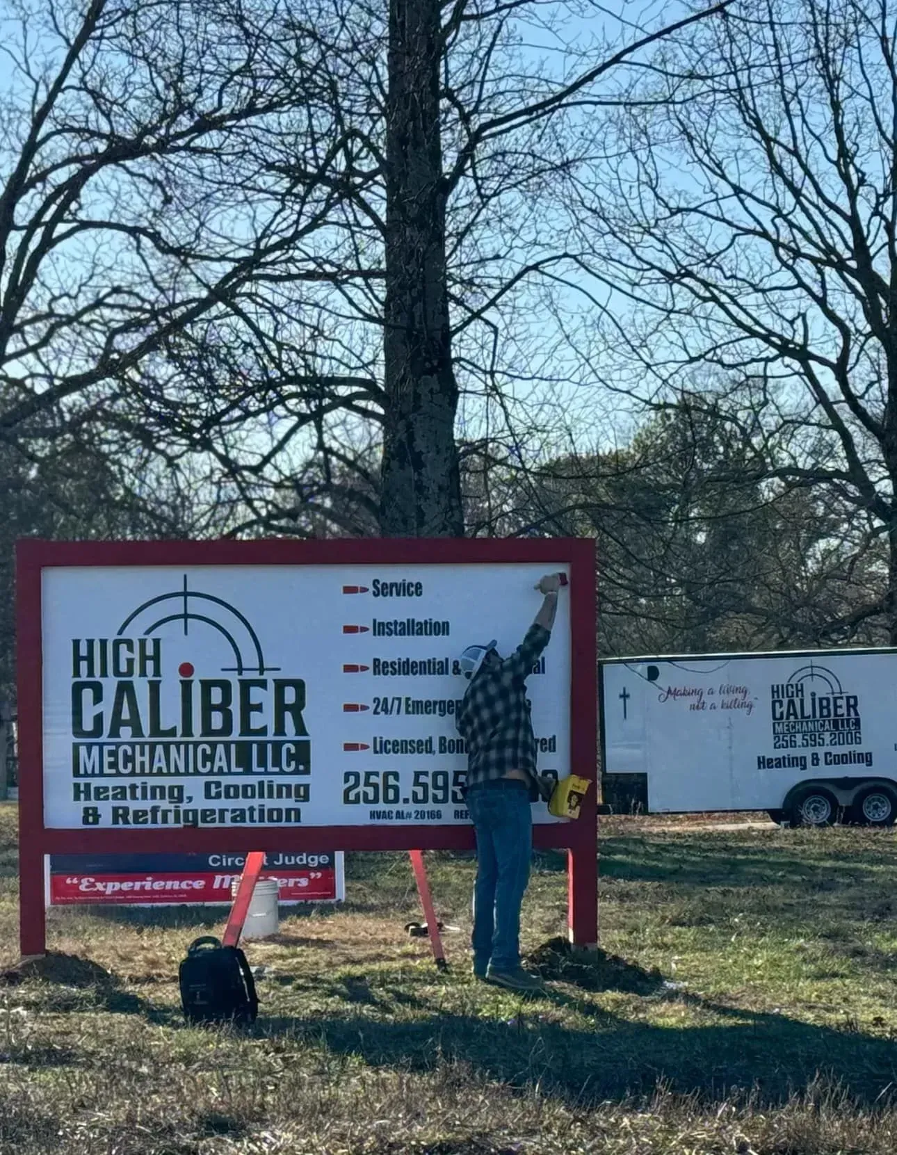 A man is standing in front of a sign that says high caliber mechanical in a field.