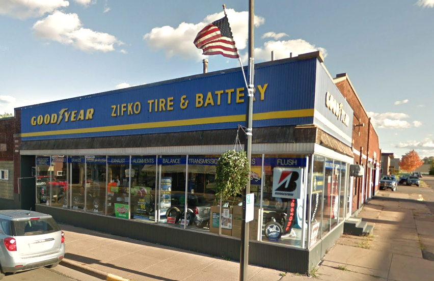 Zifko Tire & Battery shop with a blue and yellow facade, Goodyear sign, American flag, and storefront windows.