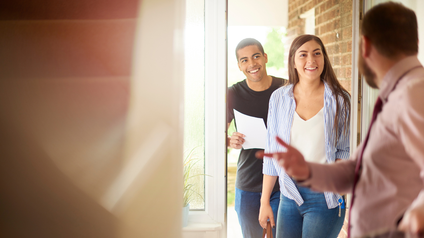 Two people at an open doorway, smiling as they greet a person inside a home.