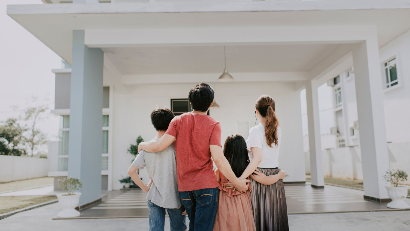 Four people stand arm-in-arm on a porch of a modern white house, viewed from behind.