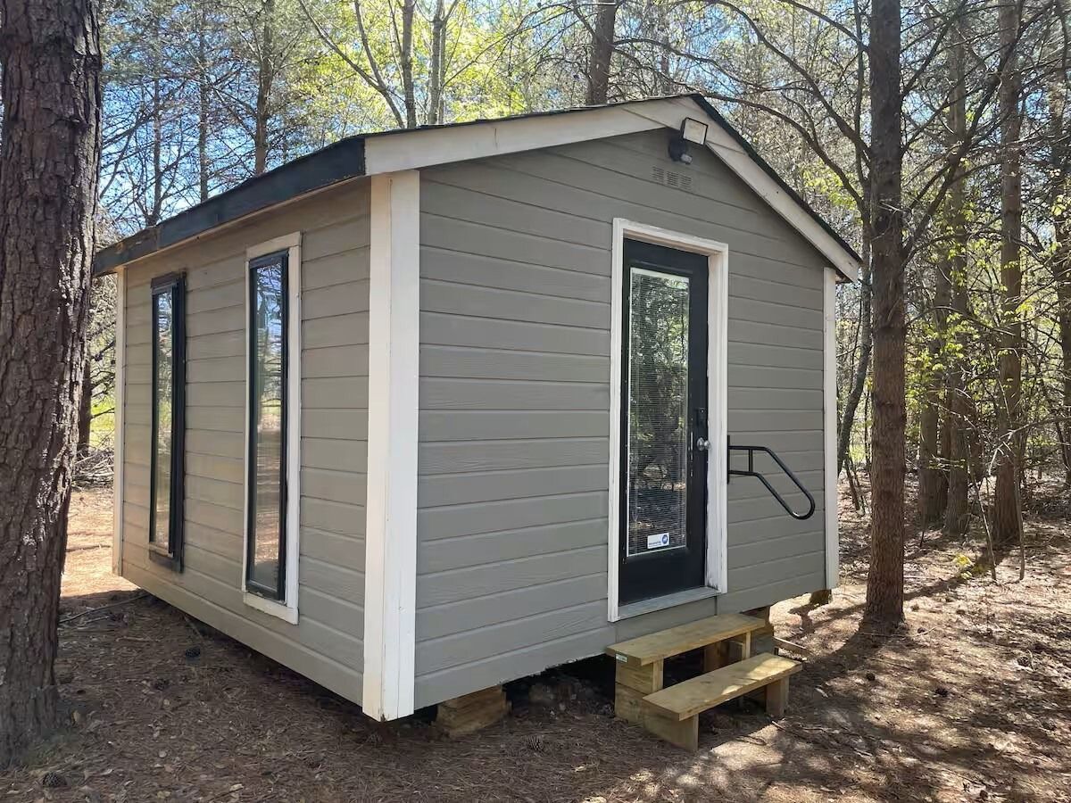 Small gray tiny house with white trim in a wooded area, with a short porch and steps.
