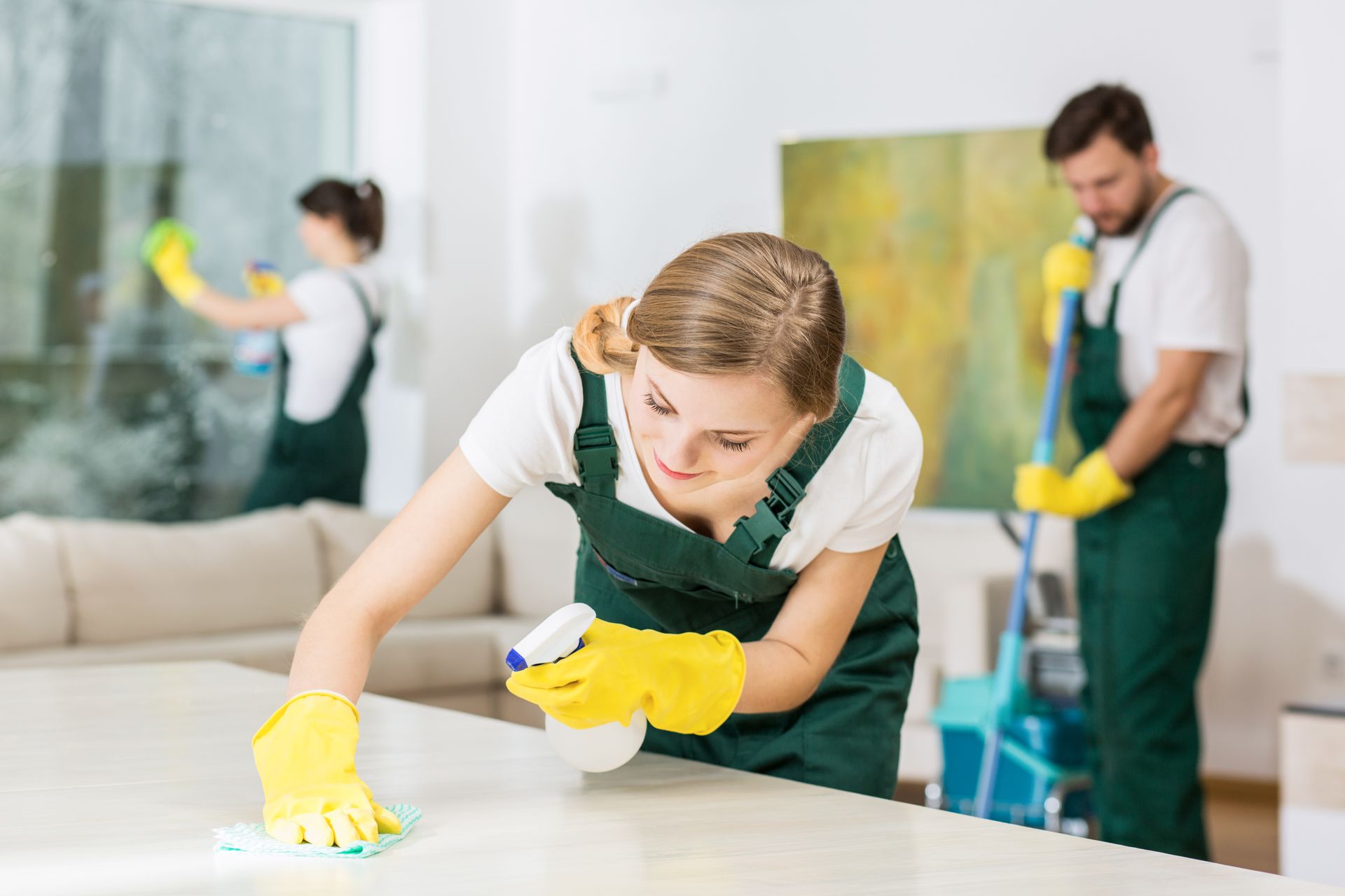 Three cleaners in green uniforms wearing yellow gloves clean a bright room with mops and cloths
