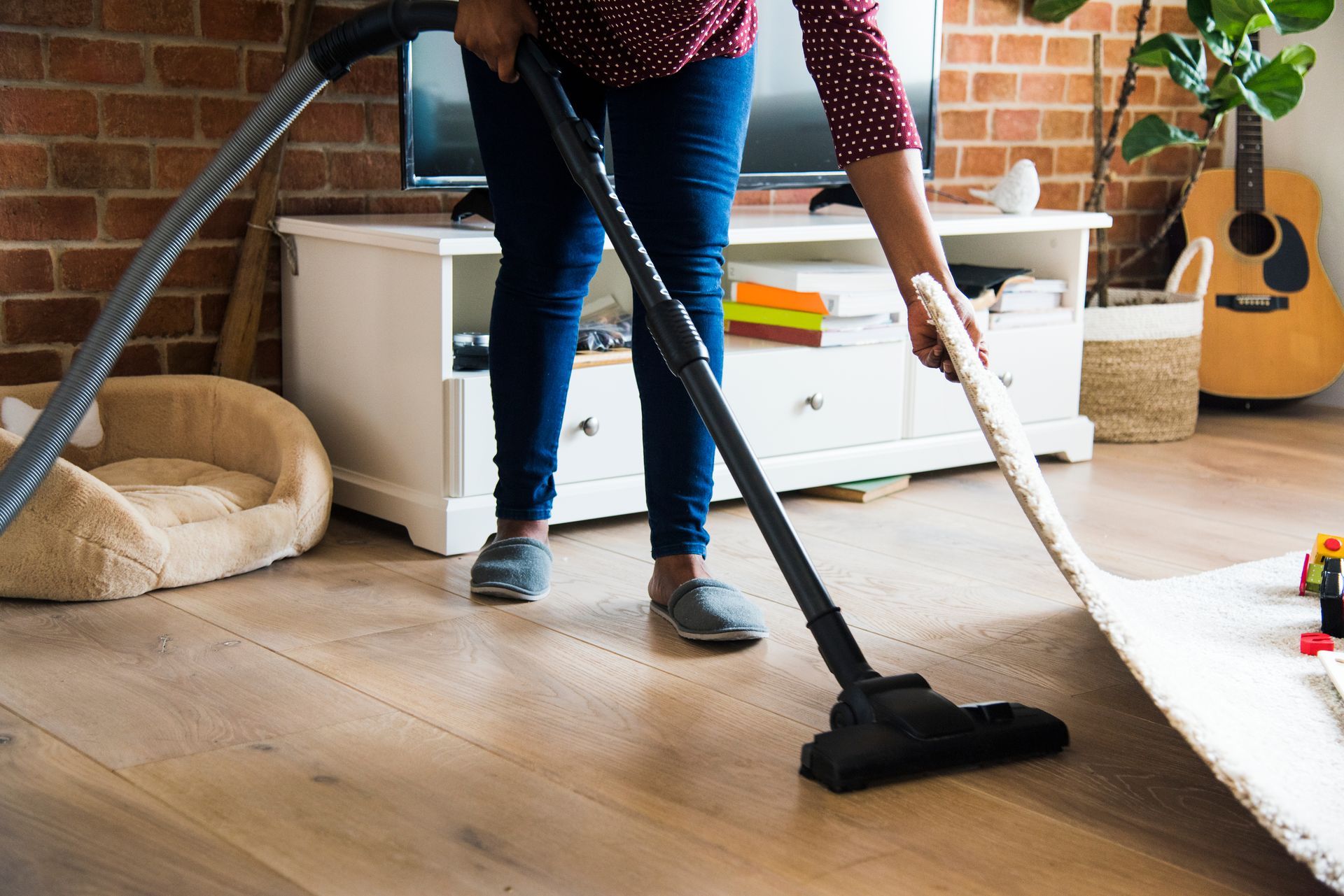 Person vacuuming a wooden floor in a bright living room with a dog bed and guitar nearby