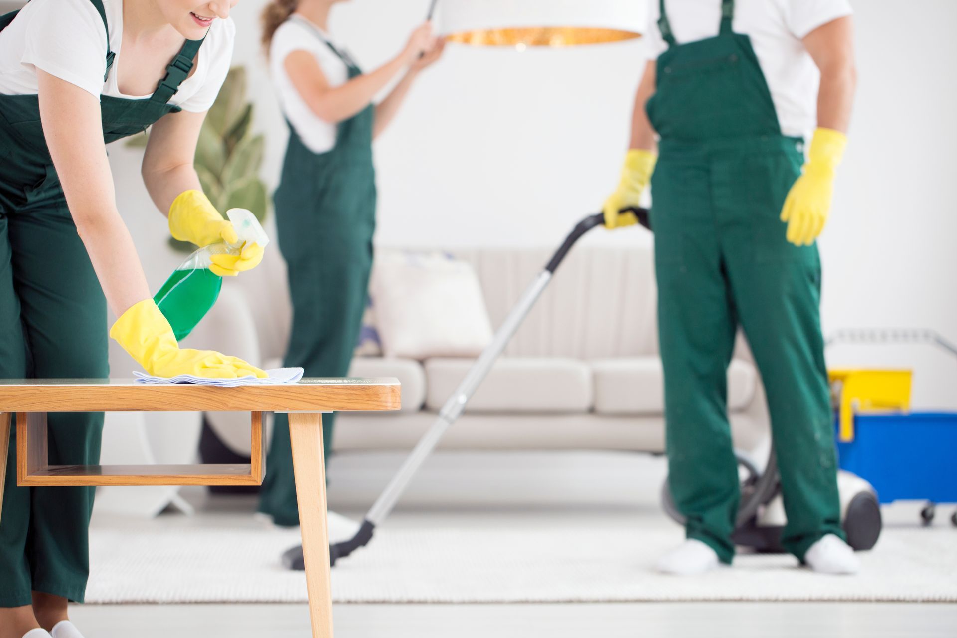 People in green aprons and yellow gloves cleaning a bright living room with a vacuum and table.