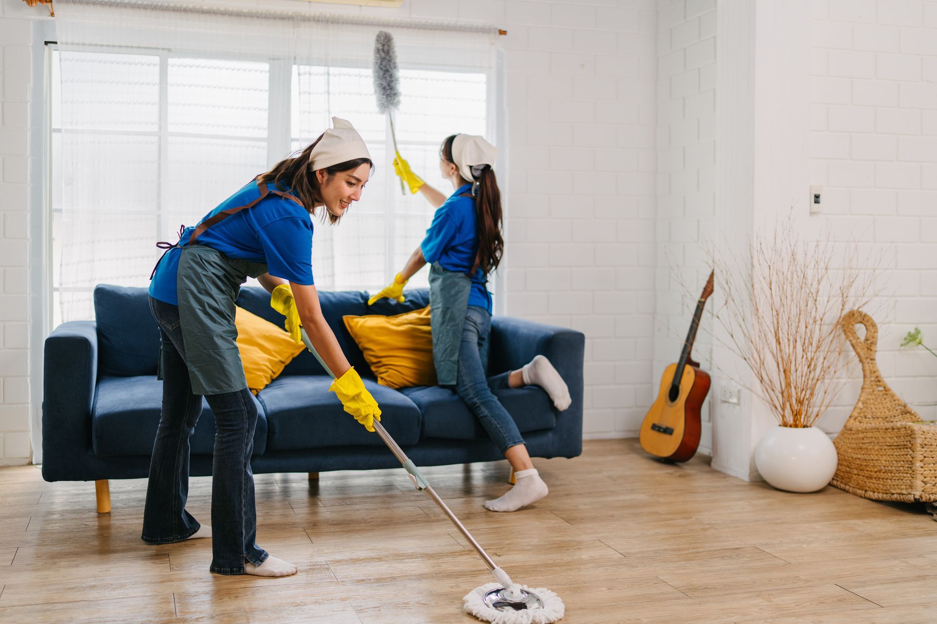 Two people cleaning a living room, mopping the floor near a blue sofa and guitar
