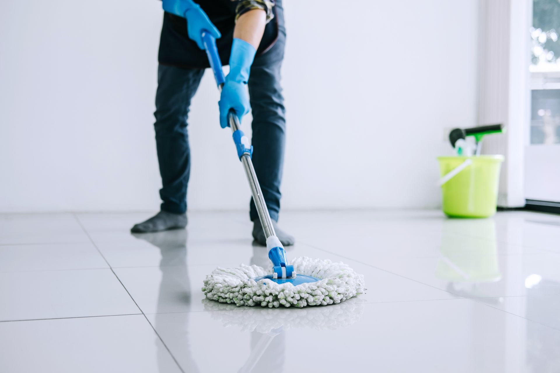 Person mopping a white tiled floor with a blue mop, cleaning solution bucket in the background
