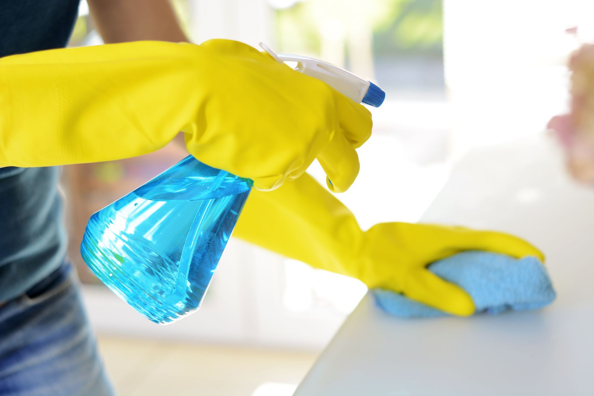 Person in yellow gloves cleaning a white surface with a blue spray bottle and cloth