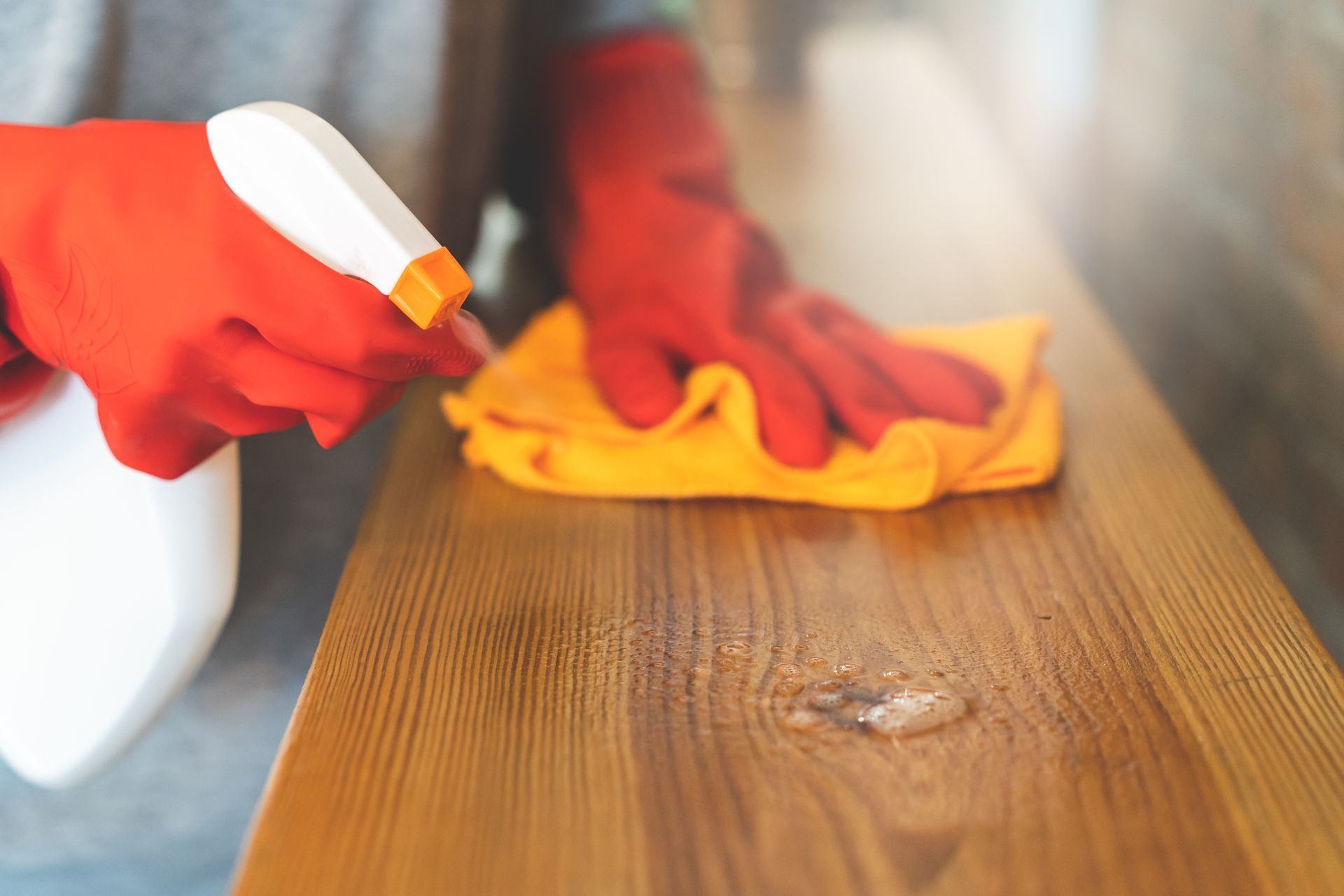 Gloved hands wiping a wooden table with a cloth and spray cleaner