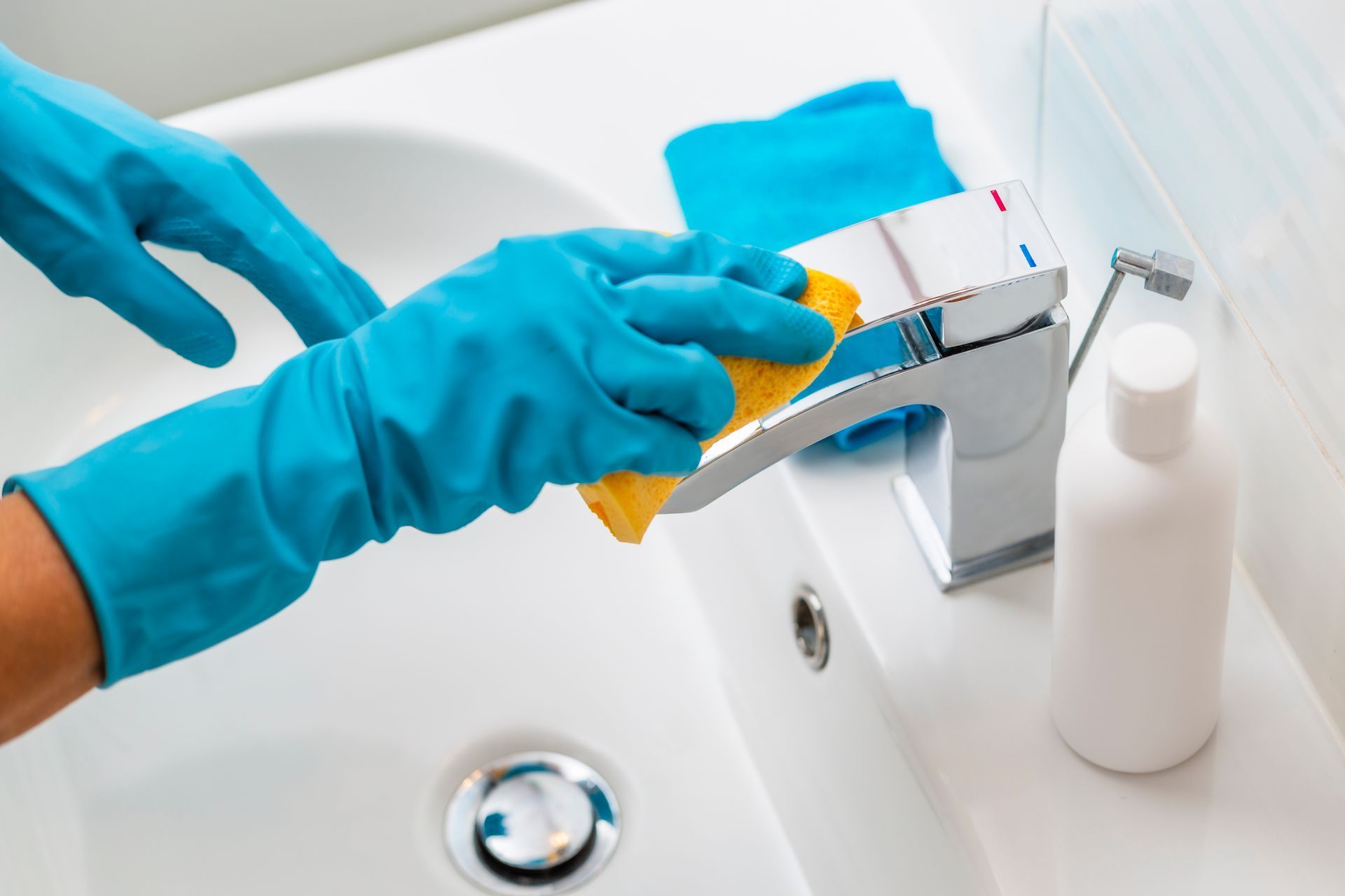 Gloved hands cleaning a bathroom sink faucet with a yellow cloth and spray bottle