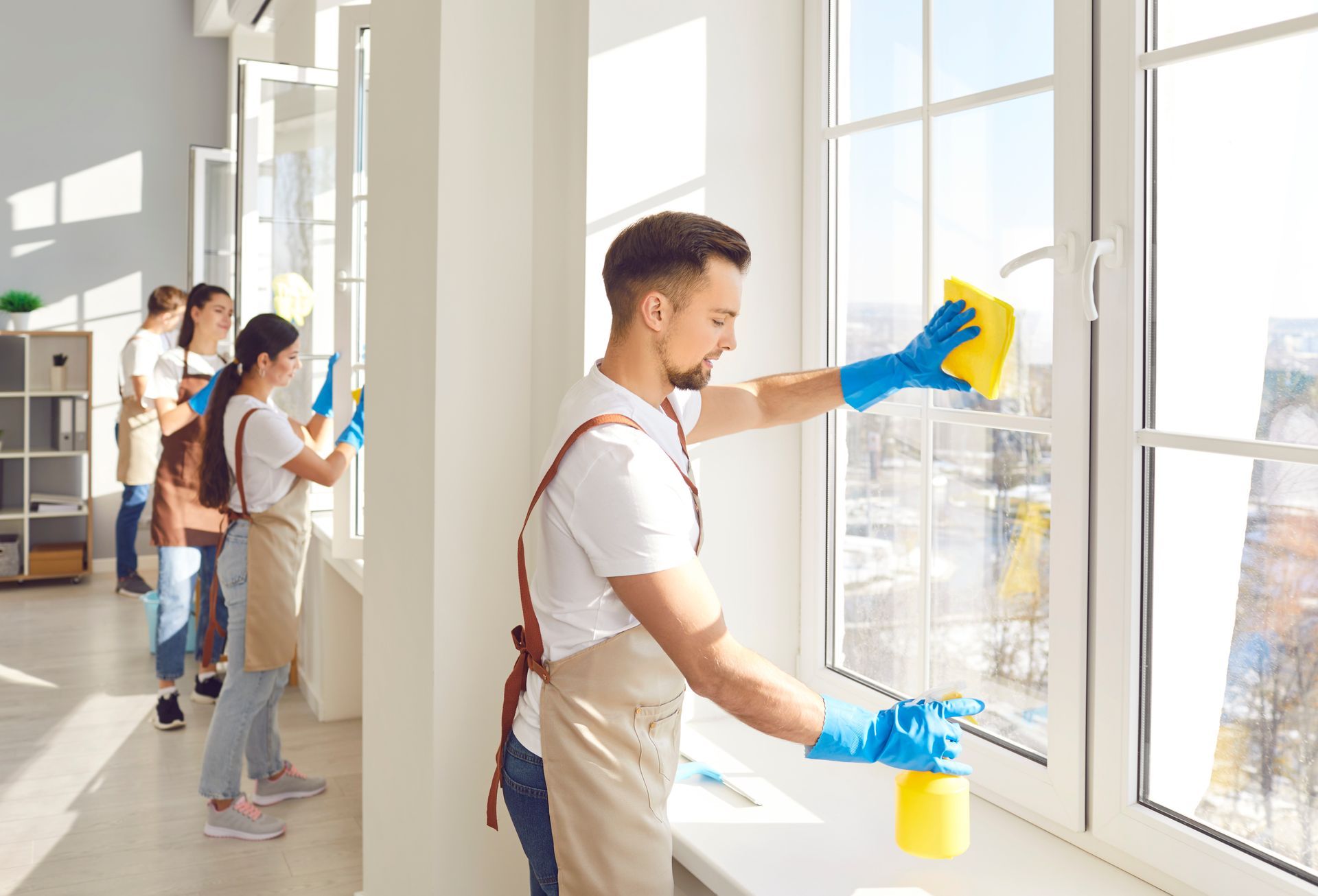 People cleaning large windows with spray bottles and cloths in a bright room