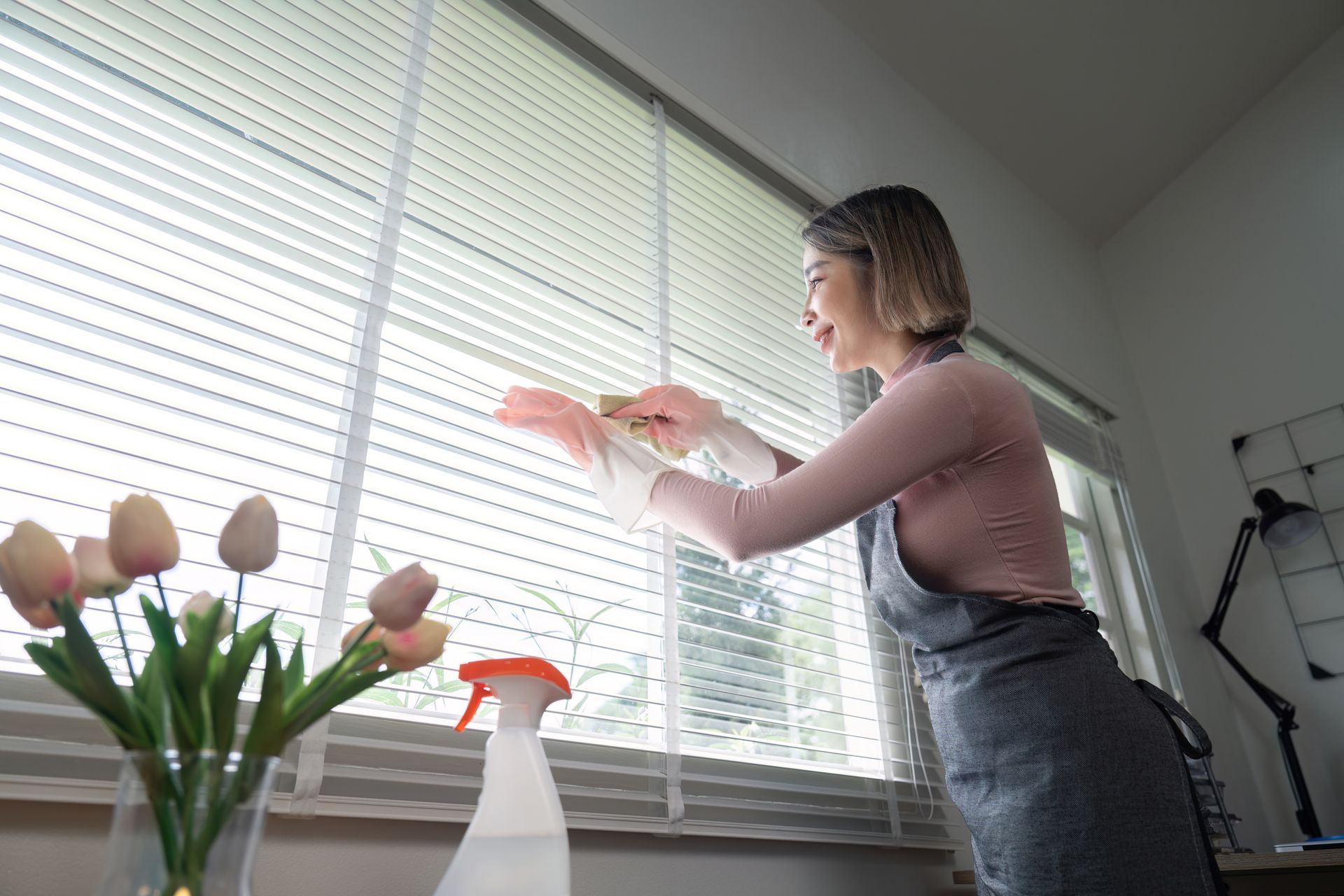 Woman cleaning window blinds beside flowers and a spray bottle on a windowsill