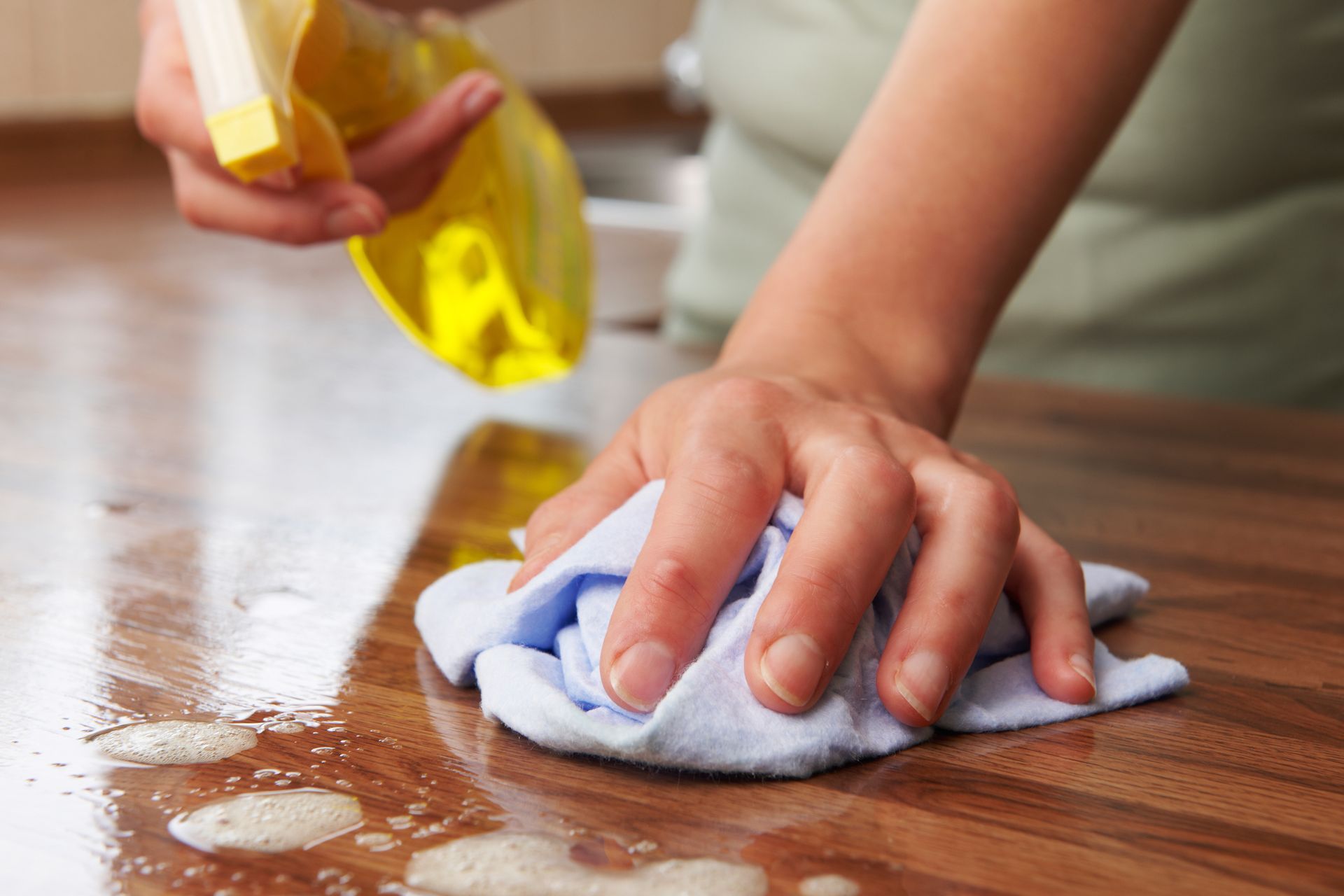 Hand wiping spilled liquid from a wooden table with a cloth, while pouring cleaner from a yellow spray bottle