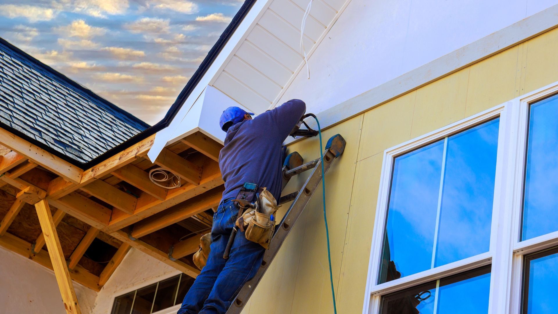Worker on a ladder repairing the exterior of a yellow house near a blue window and roofline