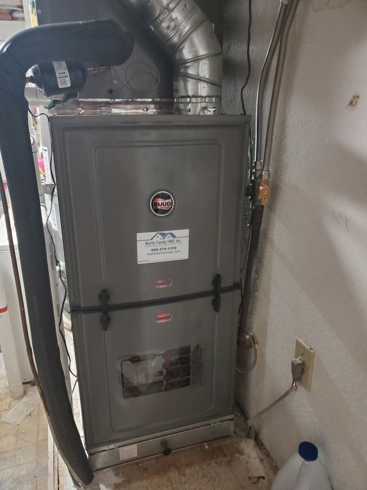 A grey, upright residential gas furnace standing in a utility area with an attached silver metal vent pipe.