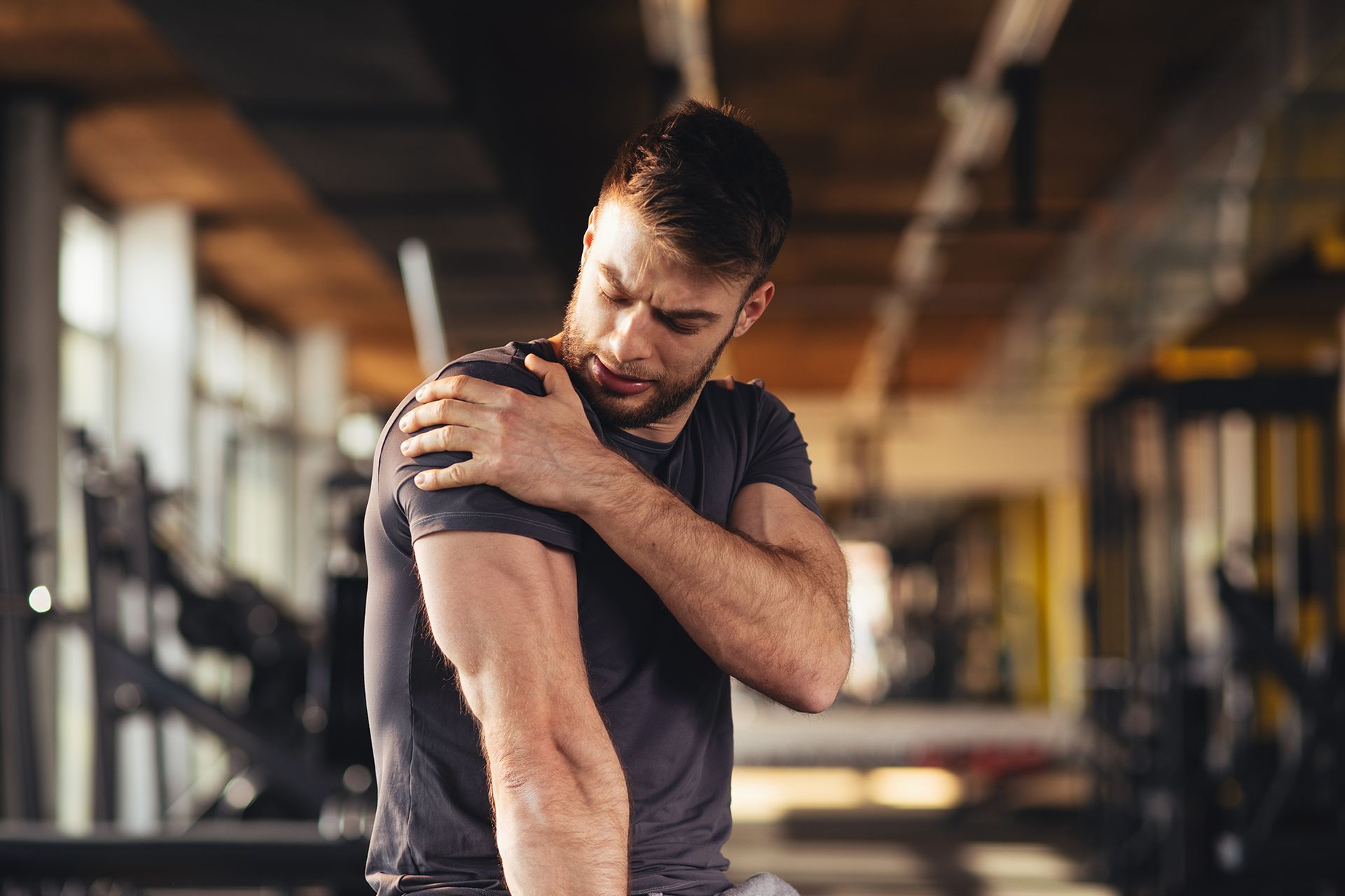 Man in a gym holding his shoulder in pain, likely from an injury. He has a grimacing expression, suggesting discomfort.