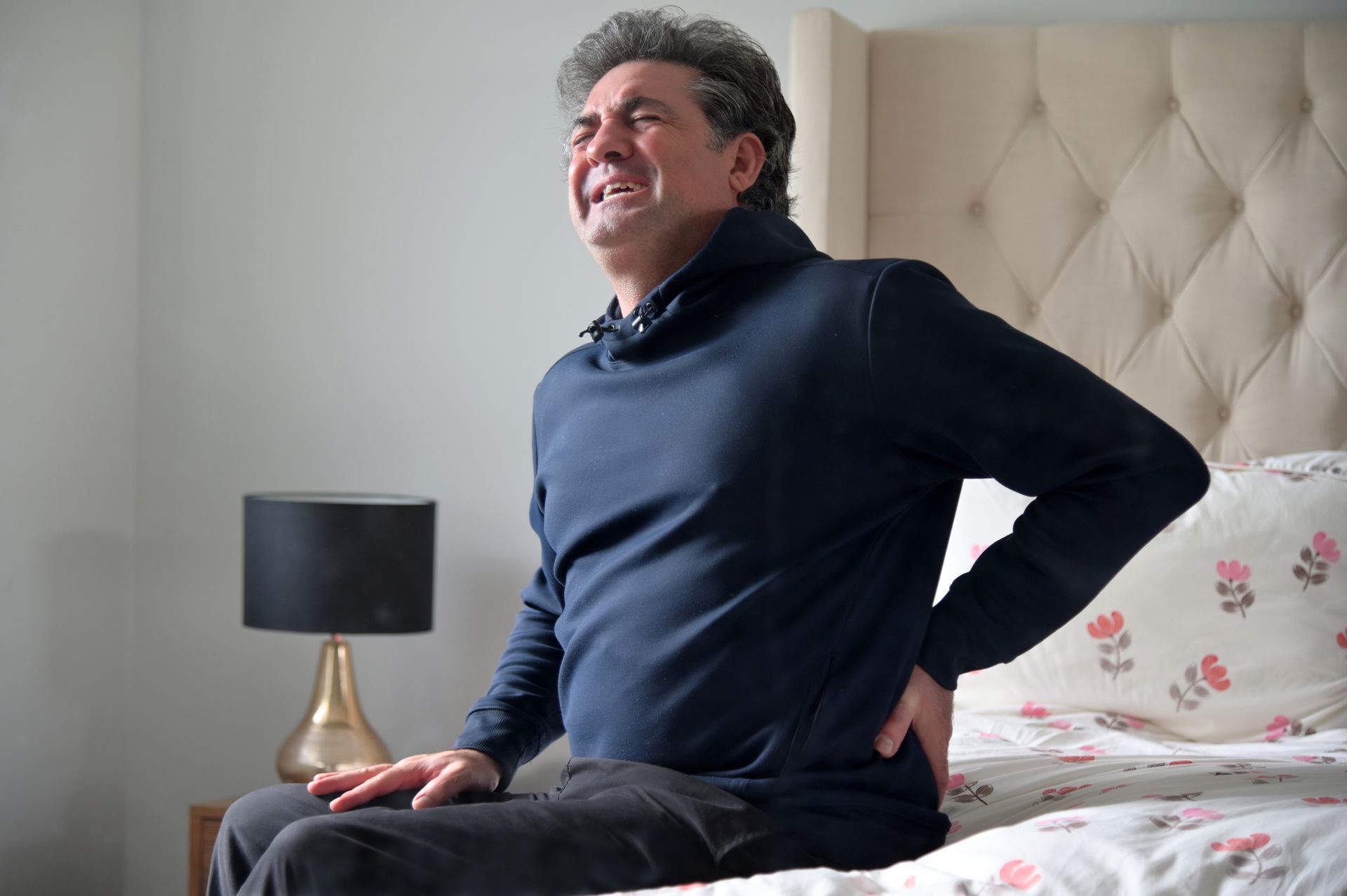 Man in dark blue sweater sitting on bed, grimacing and clutching his lower back in pain. Light-colored bedroom.