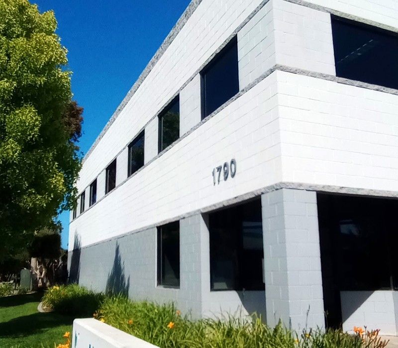 White two-story office building with dark windows and the number 1790, beside a grassy lawn and tree.