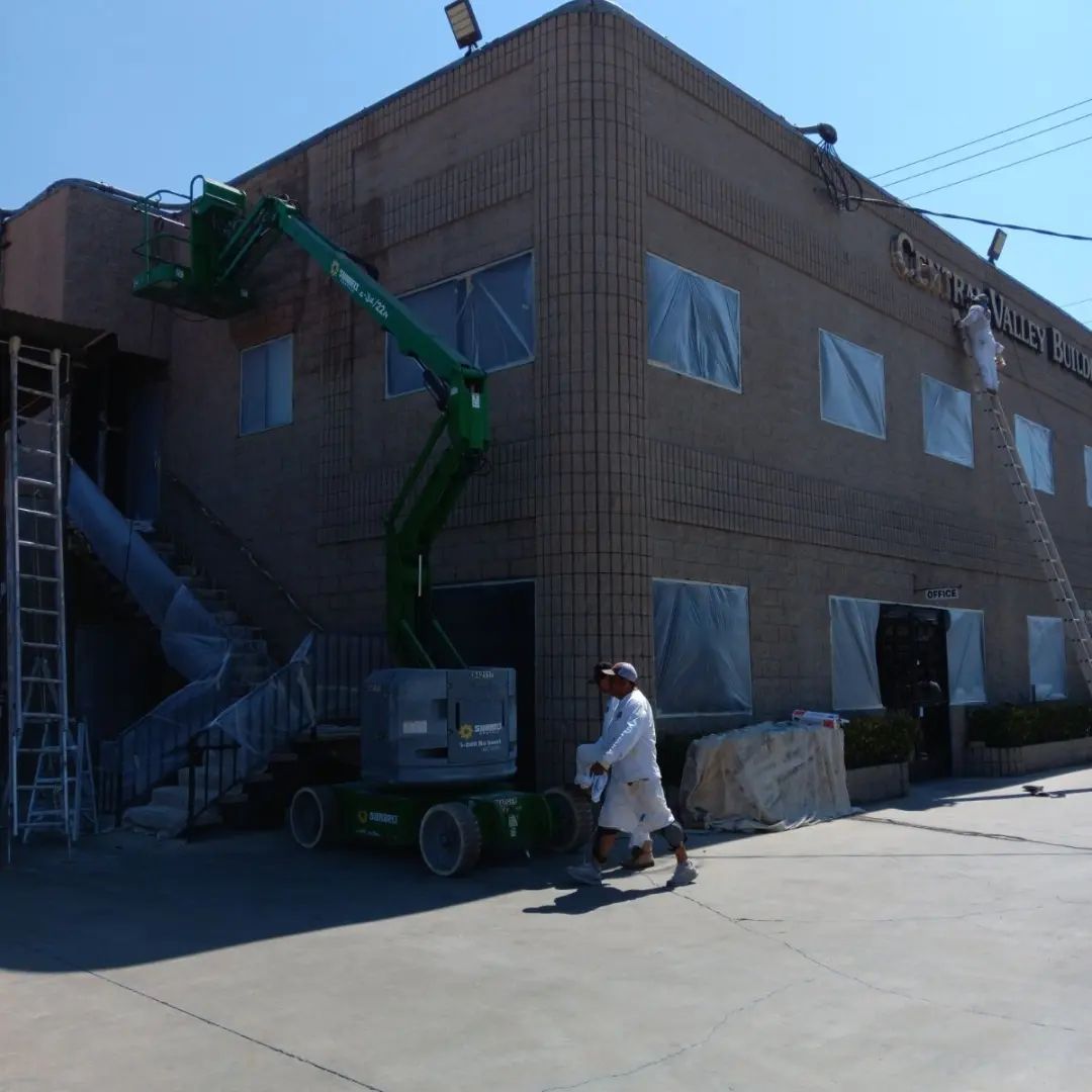 Industrial building exterior with a green boom lift and a worker on a sidewalk, covered windows visible.