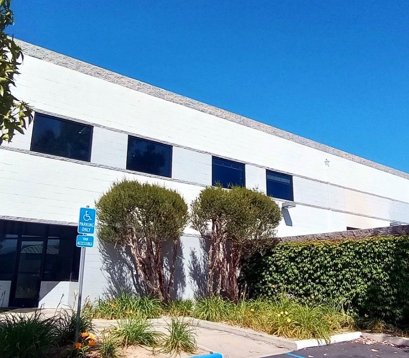 White modern office building with dark windows, blue sky, and landscaped shrubs beside the entrance