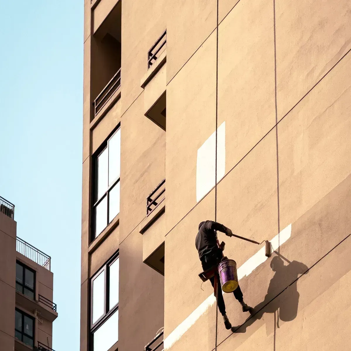 Person rappelling down a tan building wall on a rope in bright sunlight