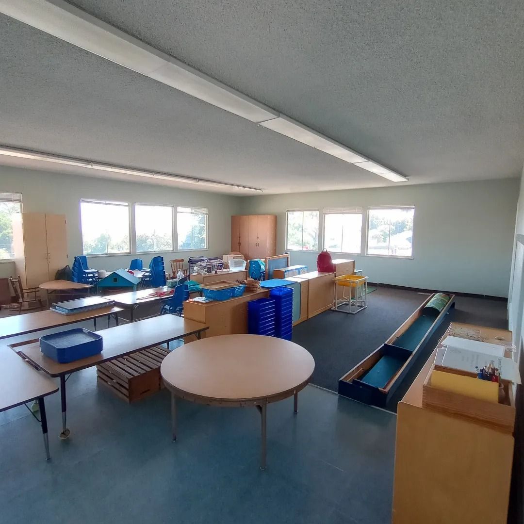 Empty classroom with tables, chairs, and storage bins under fluorescent lights.