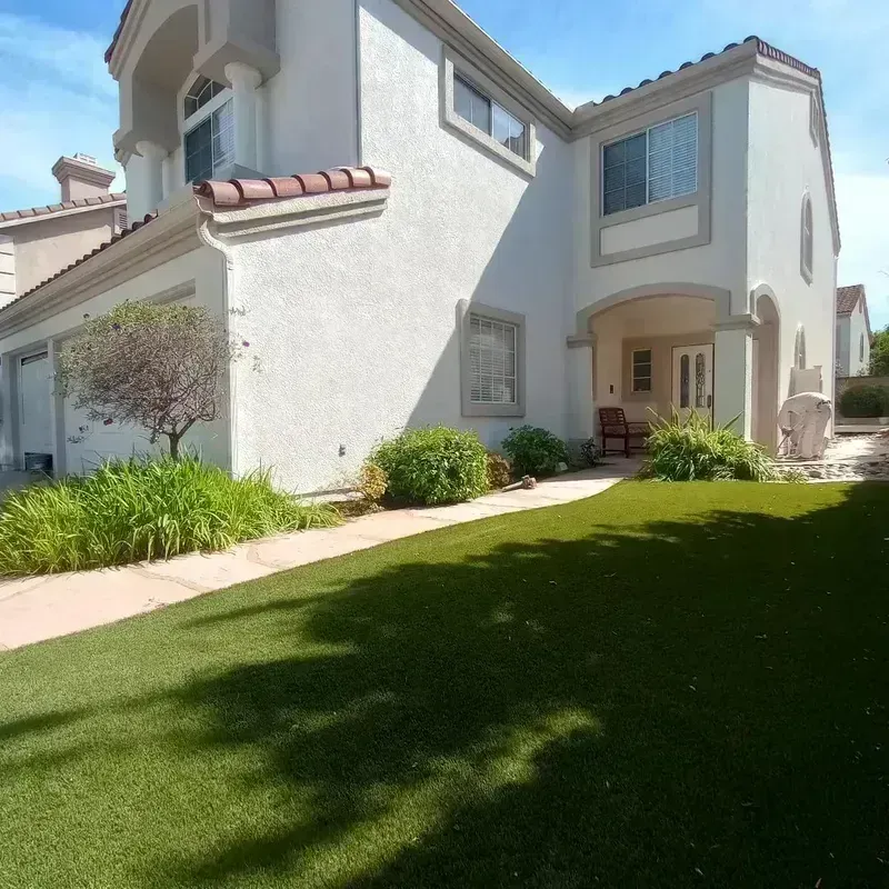 Sunlit backyard of a white stucco house with green lawn, shrubs, and a covered patio entrance