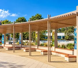 Outdoor plaza with curved wooden pergola shade, benches, trees, and blue sky